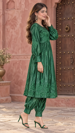 A woman models a green embroidered kurta and pant set in front of a pink stone wall and wooden door. The outfit features a long-sleeved tunic with a gathered waist and matching tapered pants.