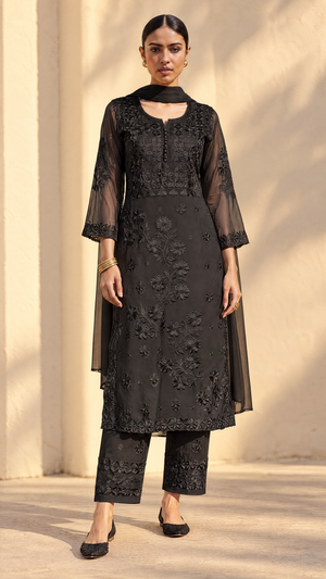 A woman models a black embroidered kurta set with matching pants and sheer long sleeves, standing against a warm beige wall with soft shadows.