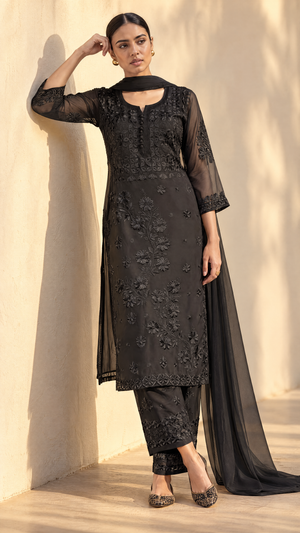 A woman models a black embroidered ethnic suit with a sheer long-sleeve kurta, matching pants, and a draped dupatta, standing against a sunlit beige wall.