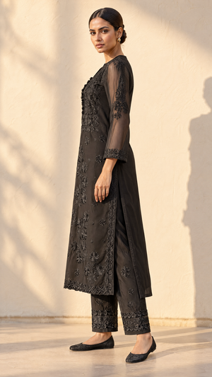 A woman modeling a black embroidered kurta and trouser set with sheer sleeves, standing against a warm beige wall with soft shadows.
