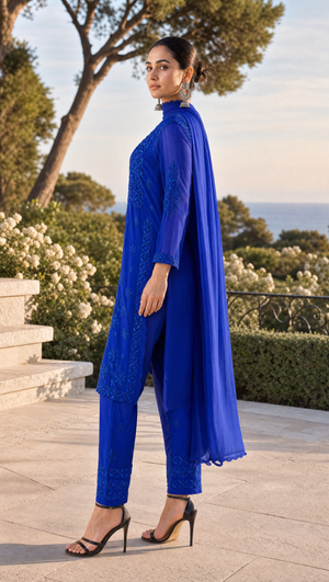 A woman modeling a royal blue ethnic three-piece suit with a long sheer kurta, matching trousers, and a draped cape, set against an outdoor garden and sea background.