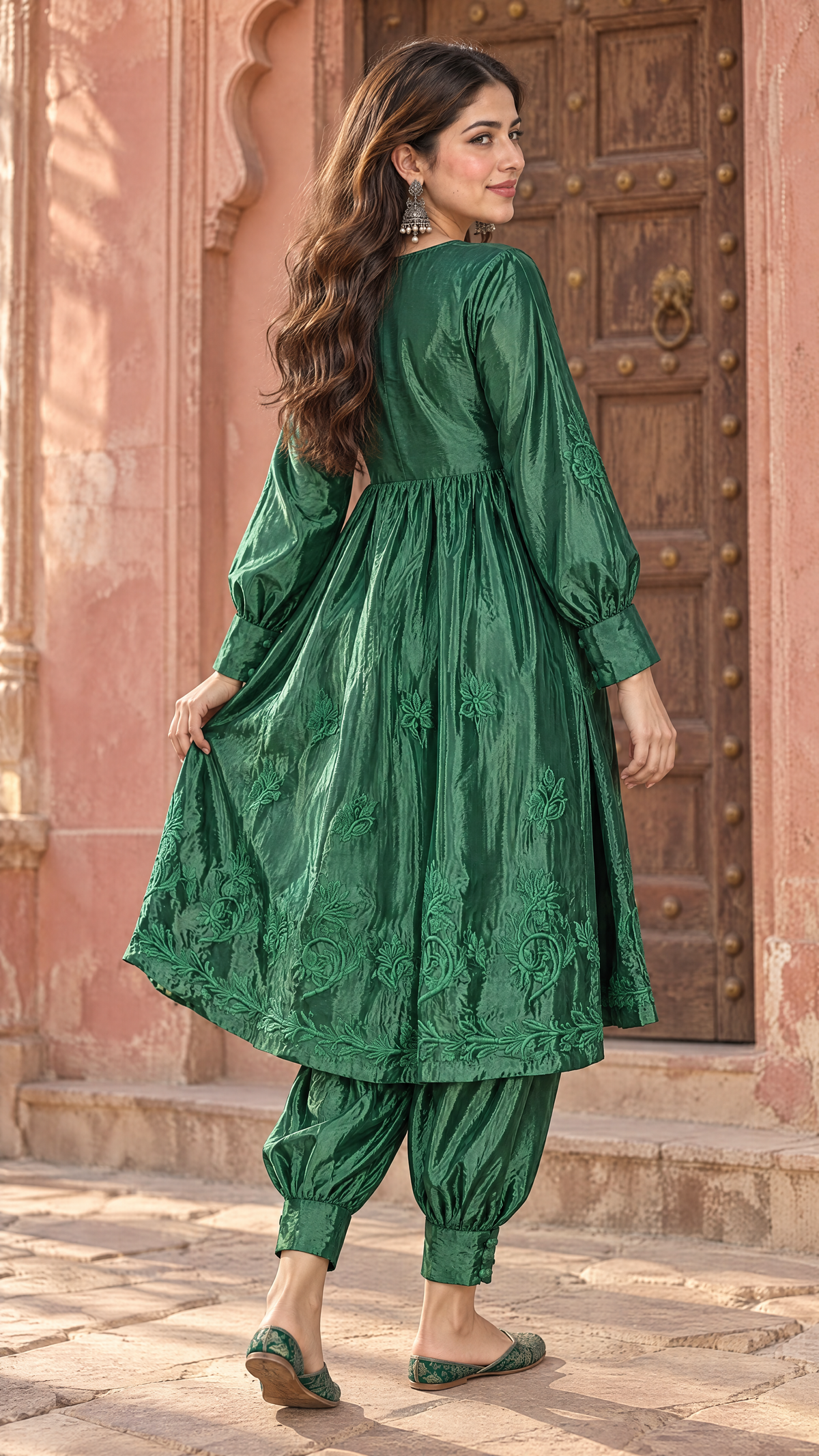 A woman wearing a green embroidered kurta and matching pants stands in front of a rustic wooden door and pink stone wall, photographed from the back and side.
