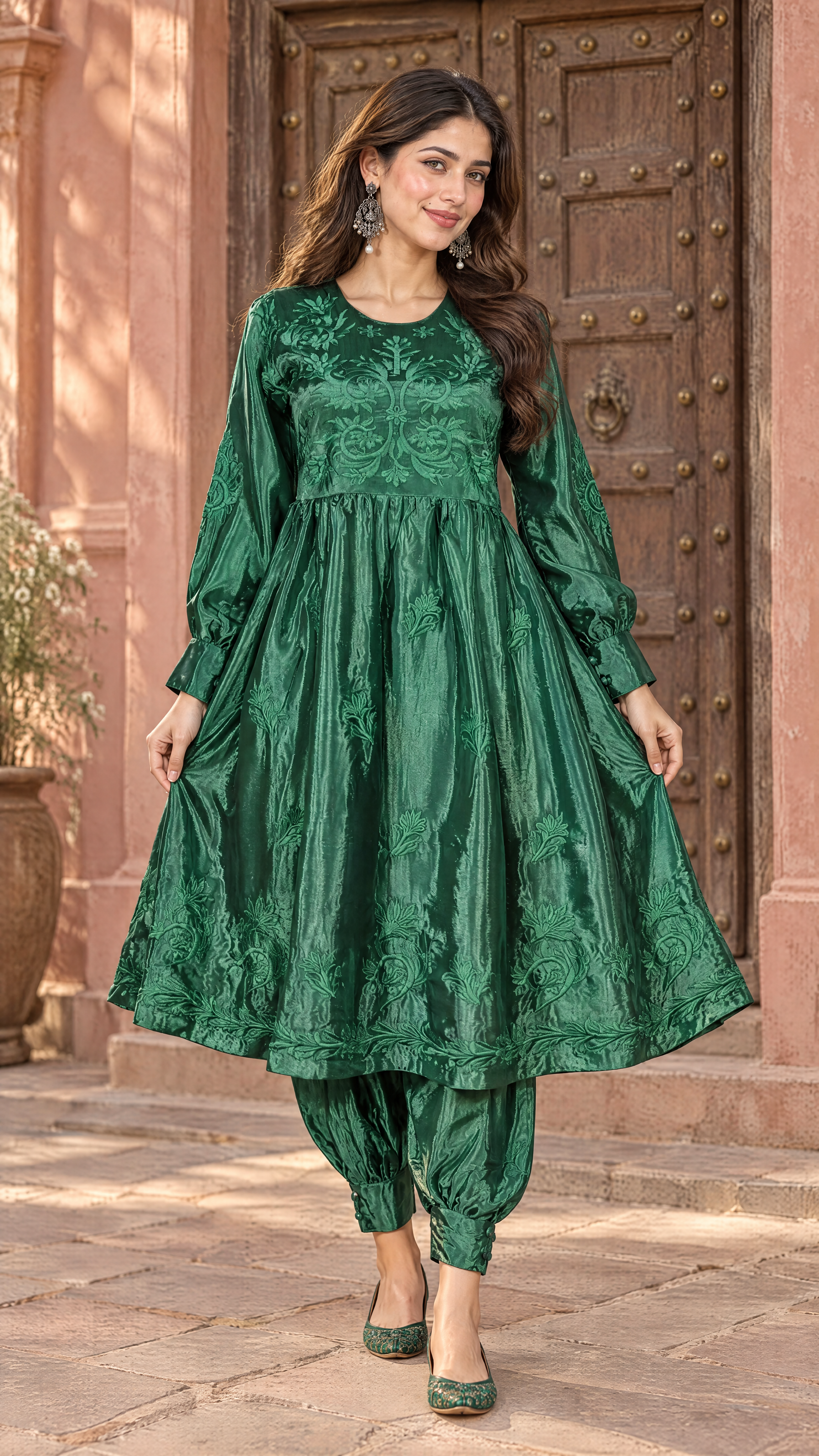 A woman models a green embroidered traditional outfit consisting of a long-sleeve kurta and matching pants, standing in front of a rustic wooden door and pink stucco wall.