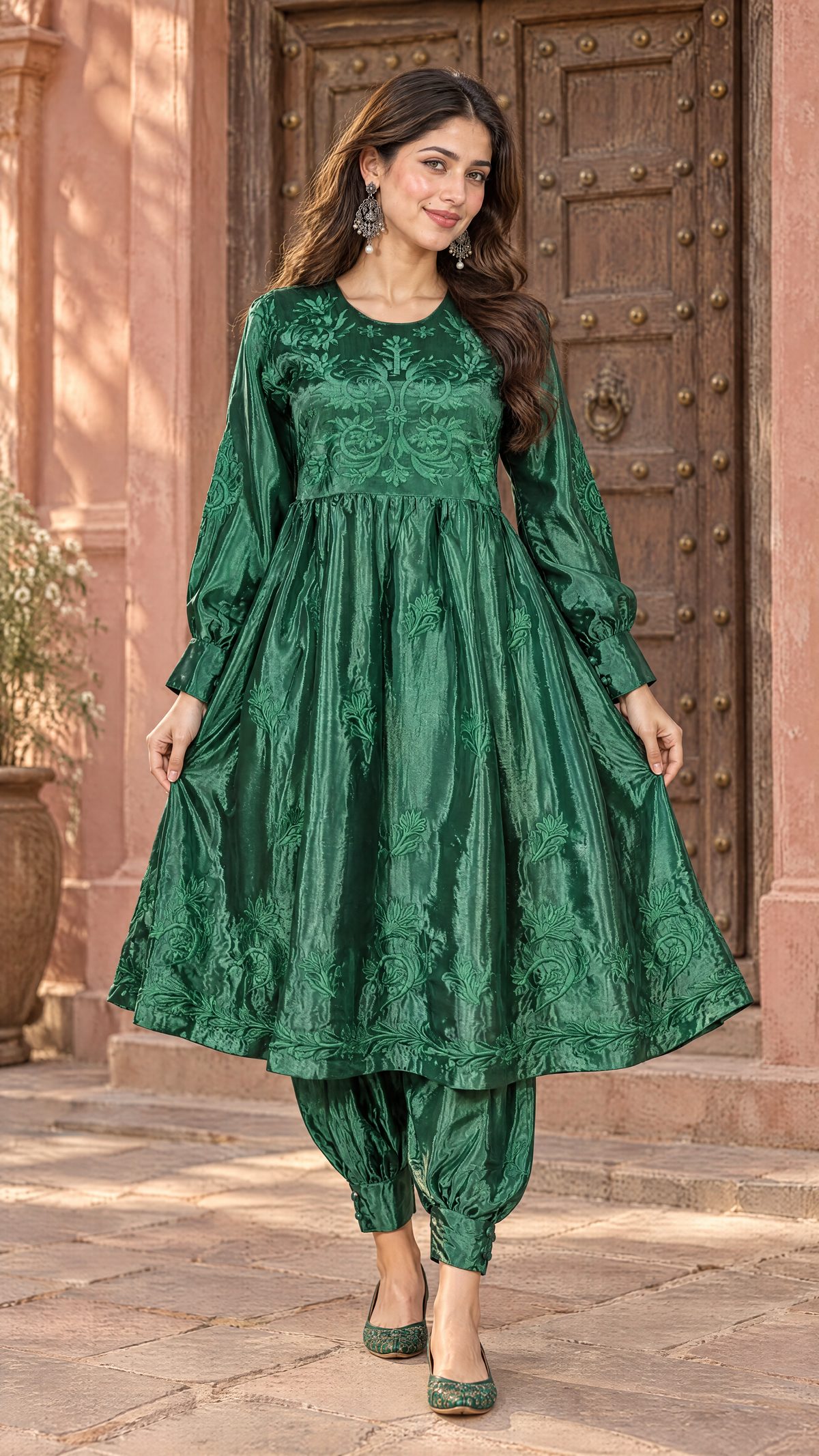 A woman models a green embroidered traditional outfit consisting of a long-sleeve kurta and matching pants, standing in front of a rustic wooden door and pink stucco wall.