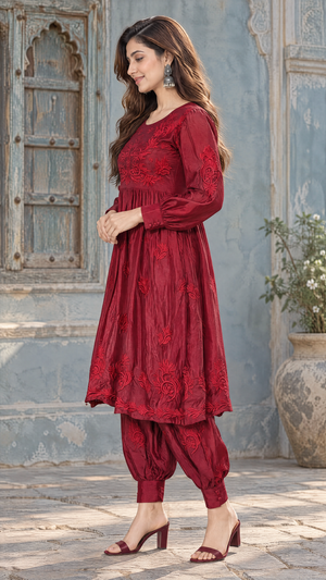 A woman models a deep red embroidered kurta set with matching pants in an outdoor courtyard setting with a rustic blue-gray wall and wooden door.