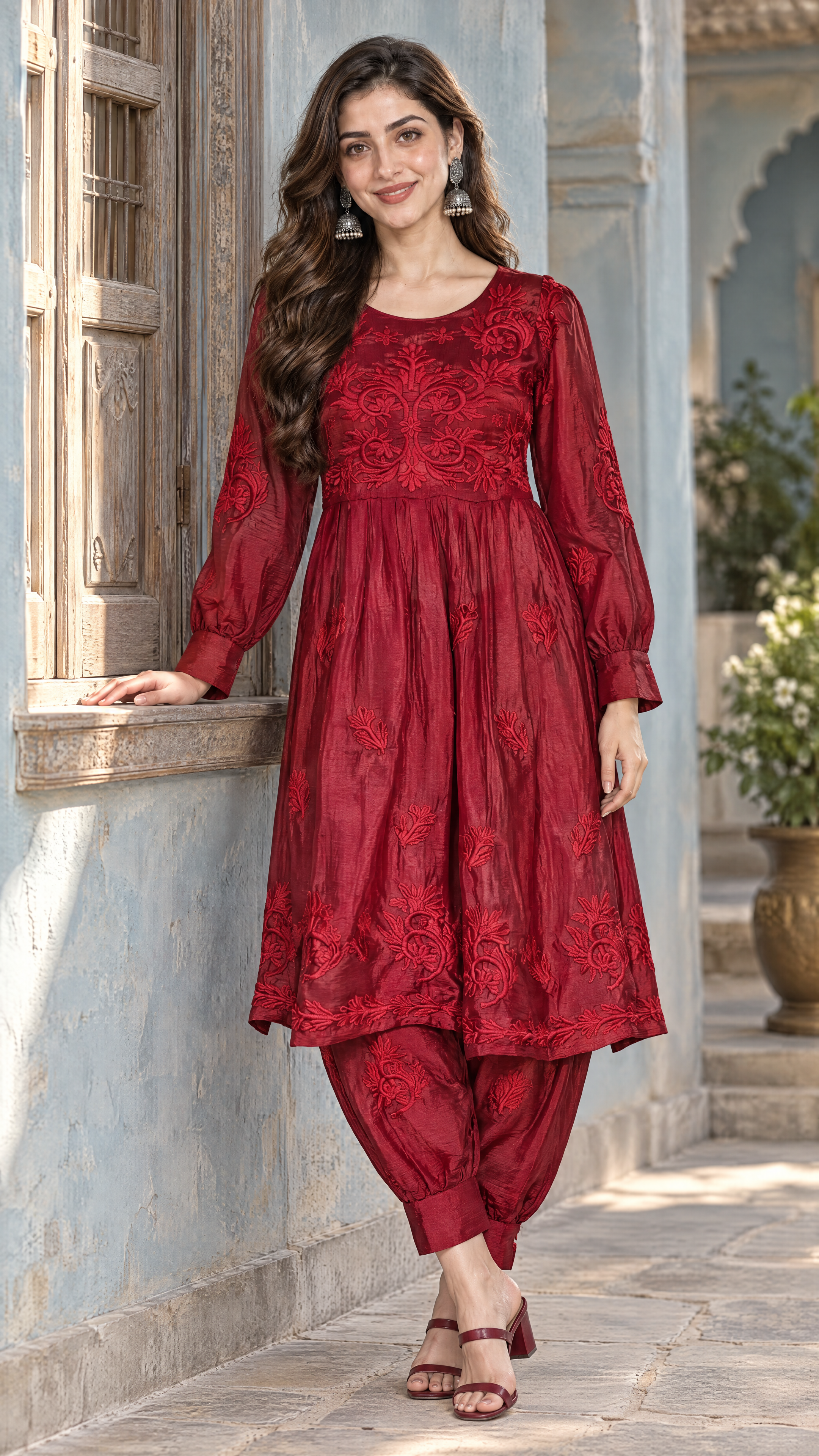 A woman models a traditional Indian salwar kameez set in deep red, featuring an embroidered long-sleeve tunic and matching patina pants, set against a light blue stone wall.