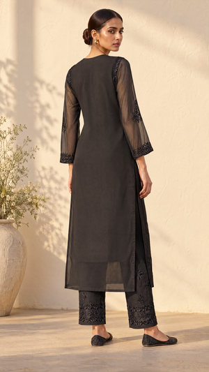 A woman modeling a black three-piece kurta set, featuring a long black kurta with sheer sleeves and black embroidered pants, standing against a warm beige wall.