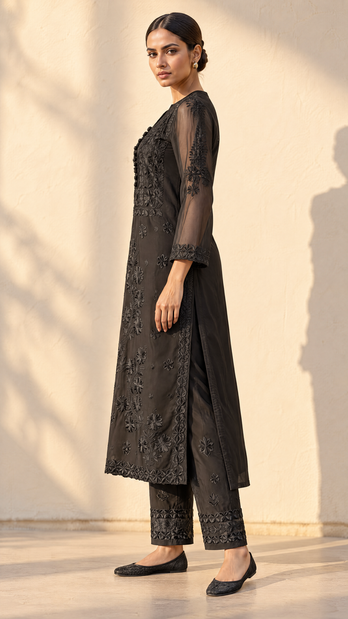A woman modeling a black embroidered kurta and trouser set with sheer sleeves, standing against a warm beige wall with soft shadows.